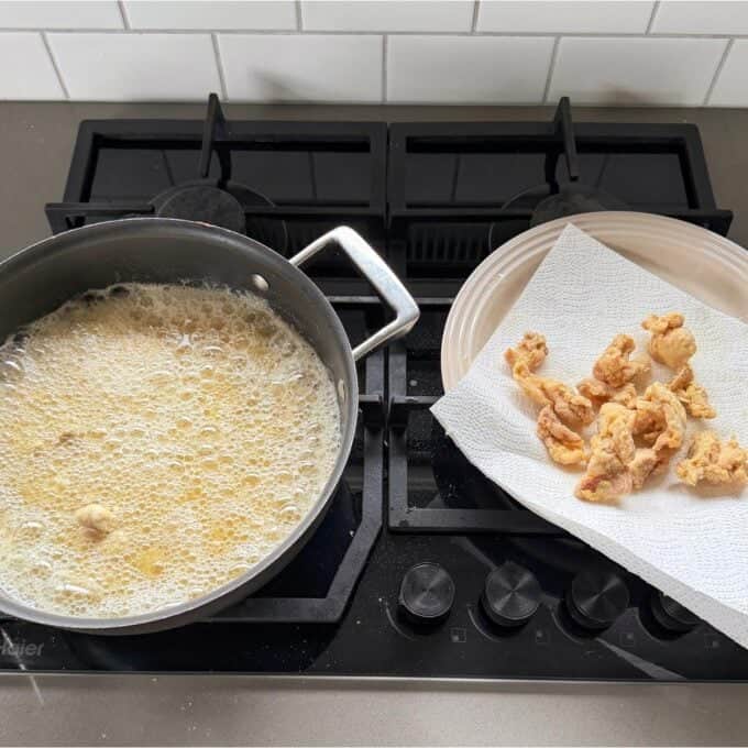 Sticky Orange Chicken Thighs A frying pan with oil and bubbling batter on a stovetop next to a plate lined with paper towels holding pieces of fried food.