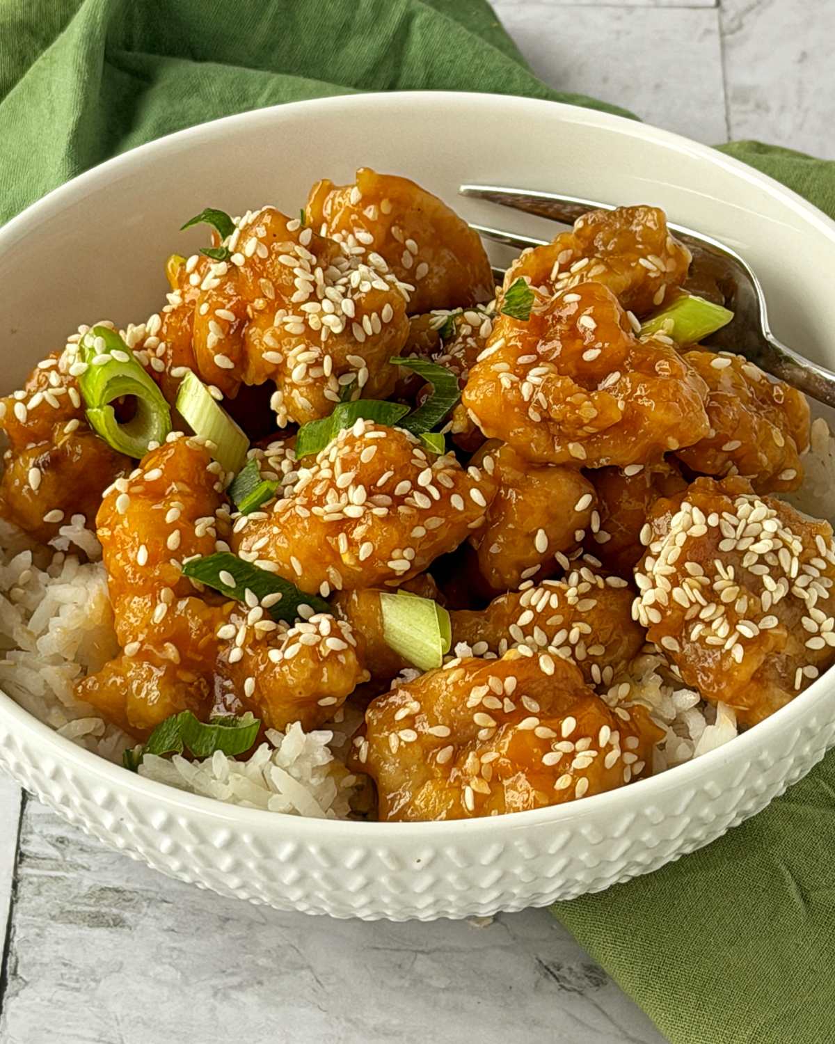 Sticky Orange Chicken Thighs A white bowl filled with rice, topped with pieces of sesame chicken, chopped green onions, and sprinkled with sesame seeds. A fork rests in the bowl.