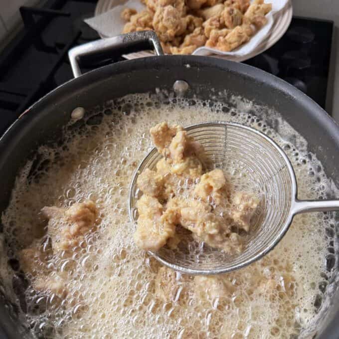 Sticky Orange Chicken Thighs Pieces of battered chicken being deep-fried in a pan, with some cooked pieces draining on a plate lined with paper towels in the background.