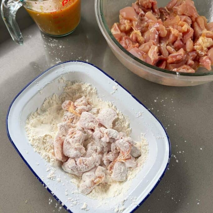 Sticky Orange Chicken Thighs Chunks of raw chicken are being coated in flour in a white dish, with a bowl of more chicken pieces and a measuring cup with liquid nearby on a countertop.