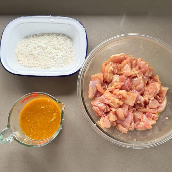 Sticky Orange Chicken Thighs A bowl of raw diced chicken, a container of flour, and a measuring cup with orange marinade are arranged on a countertop.