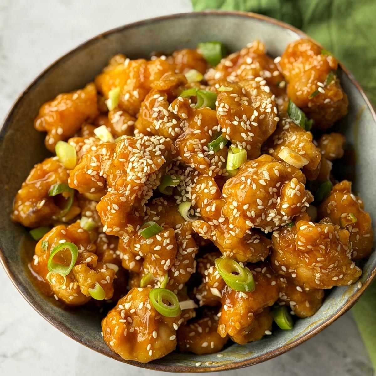 Sticky Orange Chicken Thighs A bowl of crispy fried chicken pieces coated in orange sauce, garnished with sesame seeds and sliced green onions.