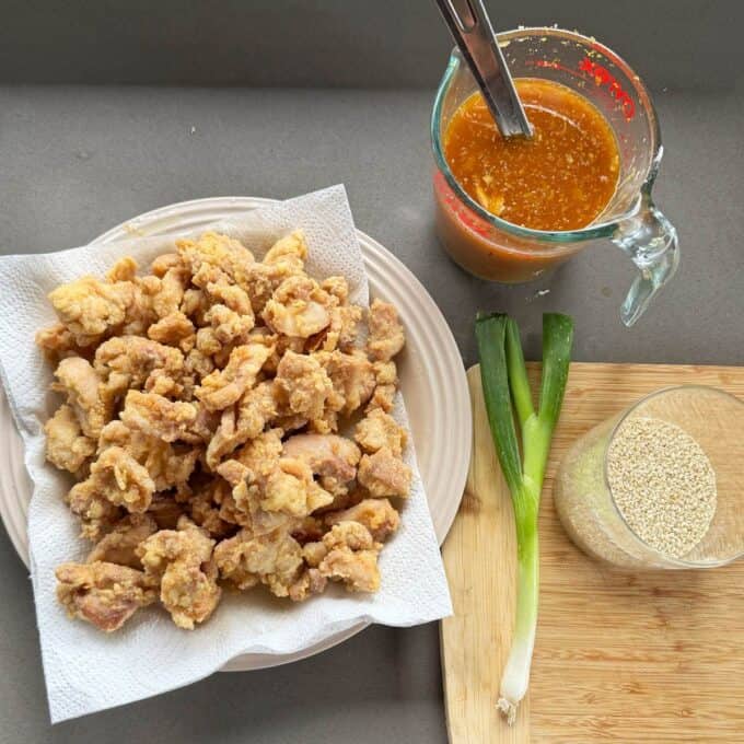 Sticky Orange Chicken Thighs Plate of fried chicken pieces on a paper towel next to a measuring cup of sauce, a green onion, and a small bowl of sesame seeds on a cutting board.