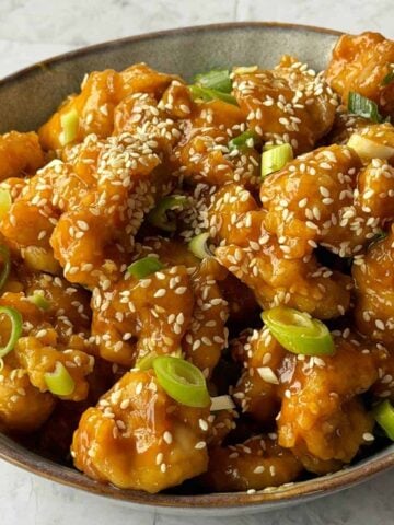 A bowl of sesame chicken pieces topped with sesame seeds and chopped green onions.