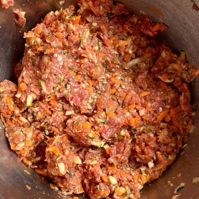 Veggie-Packed Meatballs A close-up of raw ground meat mixed with chopped vegetables and seasonings in a metal bowl.