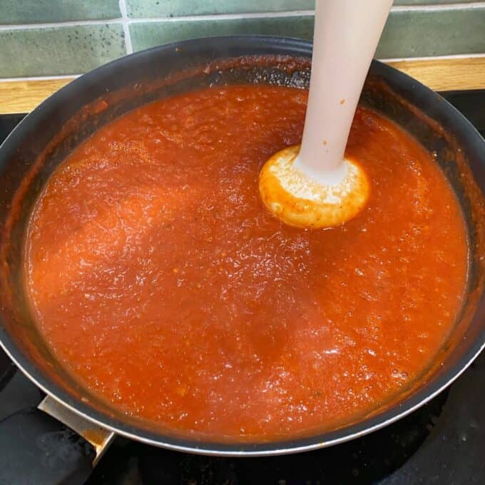 Veggie-Packed Meatballs A hand blender is blending tomato sauce in a large black pan on a stovetop, with green tiles visible in the background.