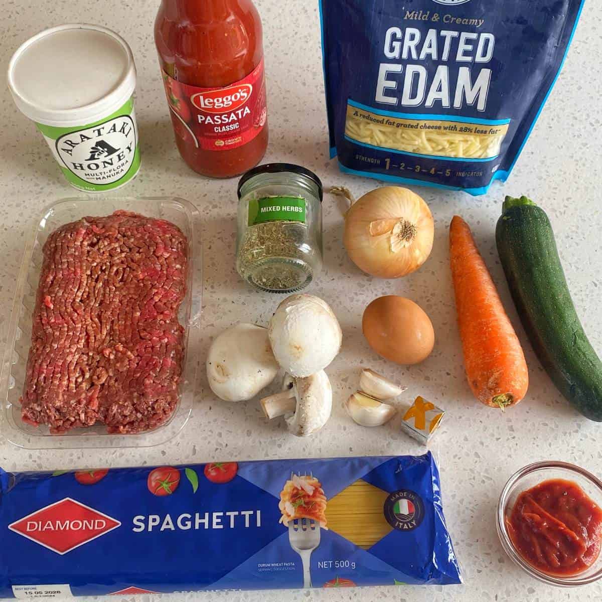 Veggie-Packed Meatballs Ingredients for spaghetti bolognese arranged on a counter, including minced meat, spaghetti, vegetables, cheese, tomato passata, honey, herbs, and tomato paste.
