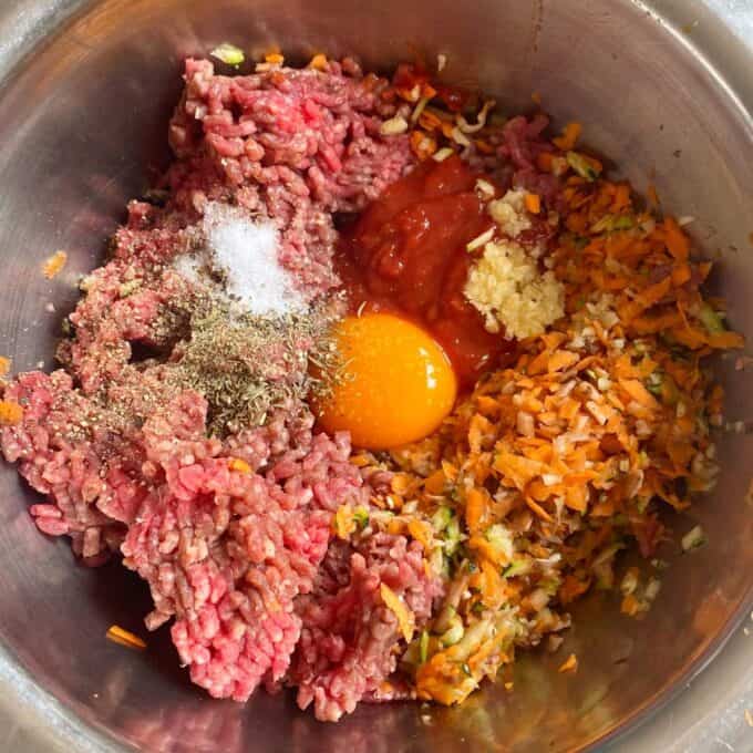 Veggie-Packed Meatballs A metal bowl containing raw ground meat, an egg yolk, shredded carrots and zucchini, tomato sauce, minced garlic, salt, and black pepper.