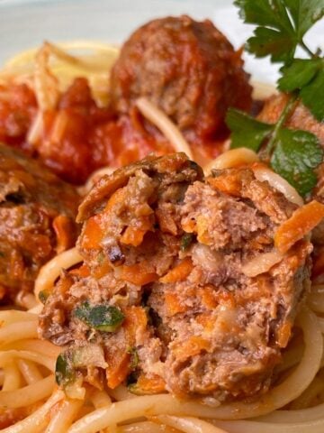 Close-up of spaghetti topped with tomato sauce and meatballs, one meatball cut open to show its vegetable filling, garnished with a parsley sprig.