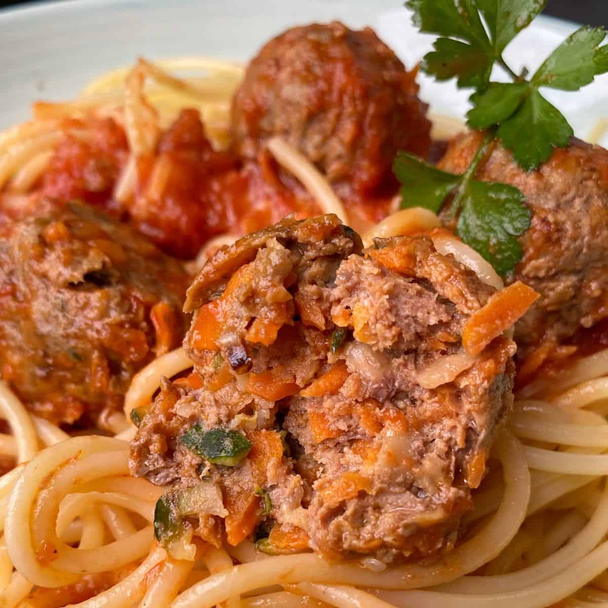 Veggie-Packed Meatballs Close-up of spaghetti topped with tomato sauce and meatballs, one meatball cut open to show its vegetable filling, garnished with a parsley sprig.