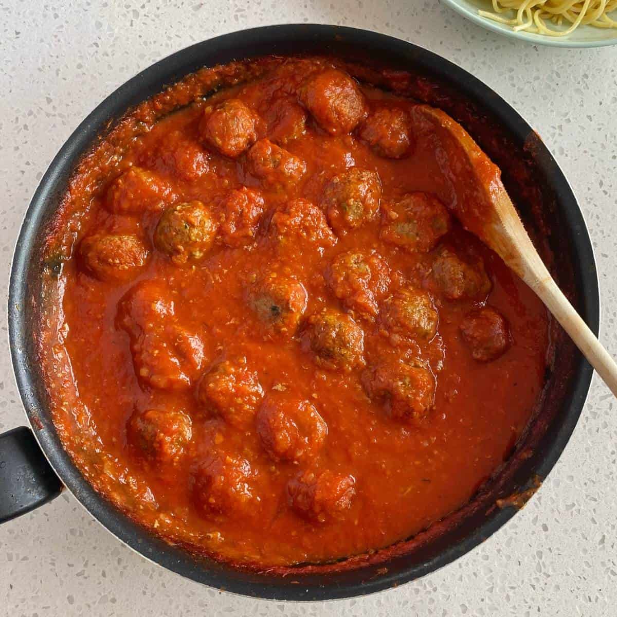 Veggie-Packed Meatballs A pan filled with meatballs in tomato sauce, with a wooden spoon resting on the edge and some spaghetti visible on a plate in the corner.