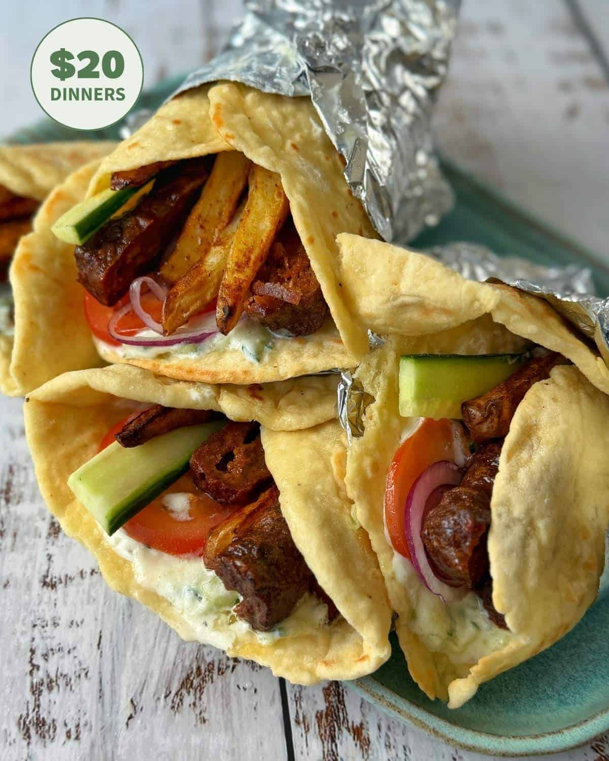 Three pita wraps filled with fries, tomatoes, cucumbers, red onions, and sauce, partially wrapped in foil, are arranged on a light blue plate. A $20 dinners label is visible in the corner.