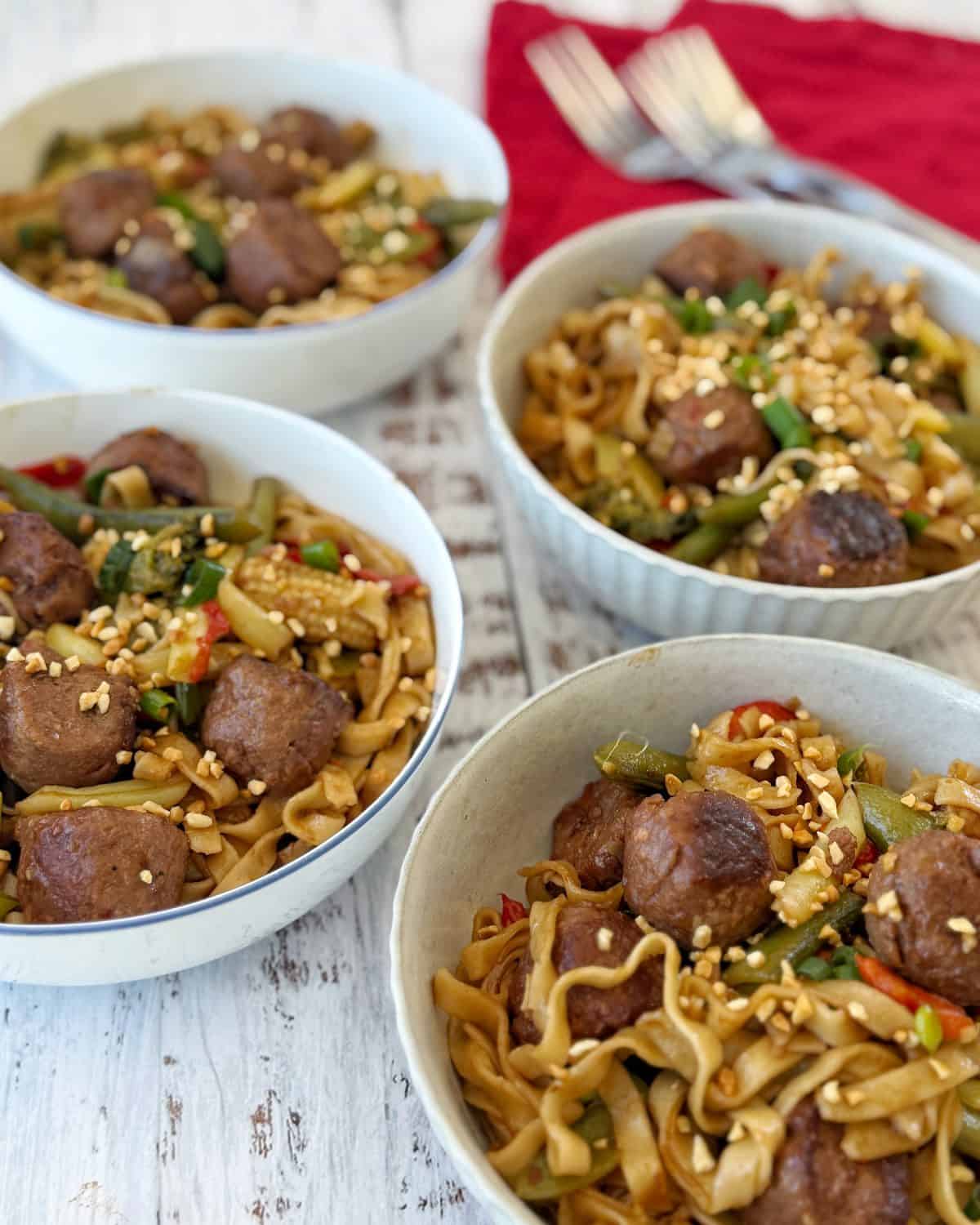 Meatball Noodle Stir Fry Four bowls of noodle stir-fry with meatballs, vegetables, and sesame seeds are placed on a white wooden surface next to a red napkin with forks.