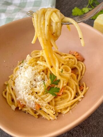 Bowl of Spaghetti Carbonara topped with grated cheese and parsley, with a fork lifting a portion; wedge of cheese and a checked cloth in the background.
