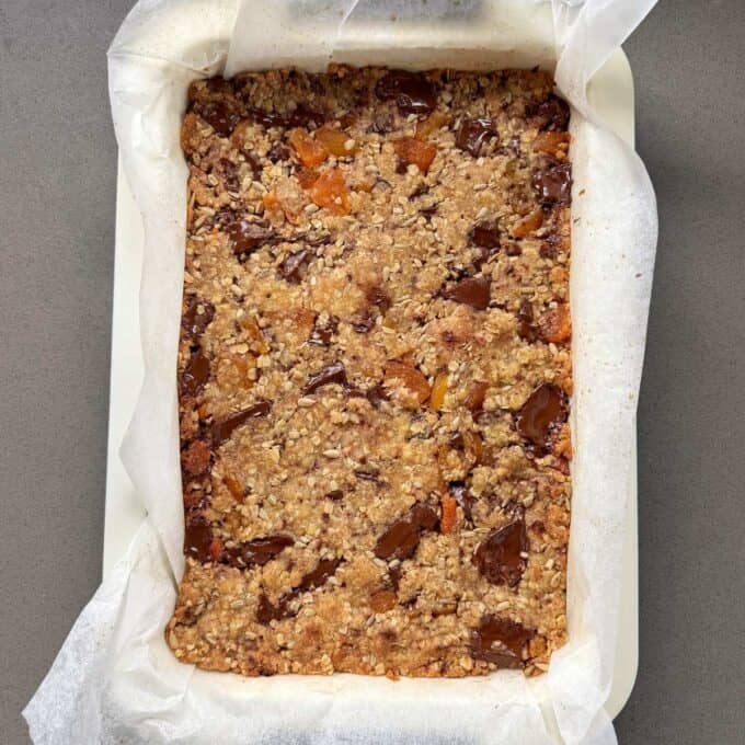 Apricot Bumper Bars A tray of baked oat and chocolate chip granola bars lined with parchment paper on a gray surface.