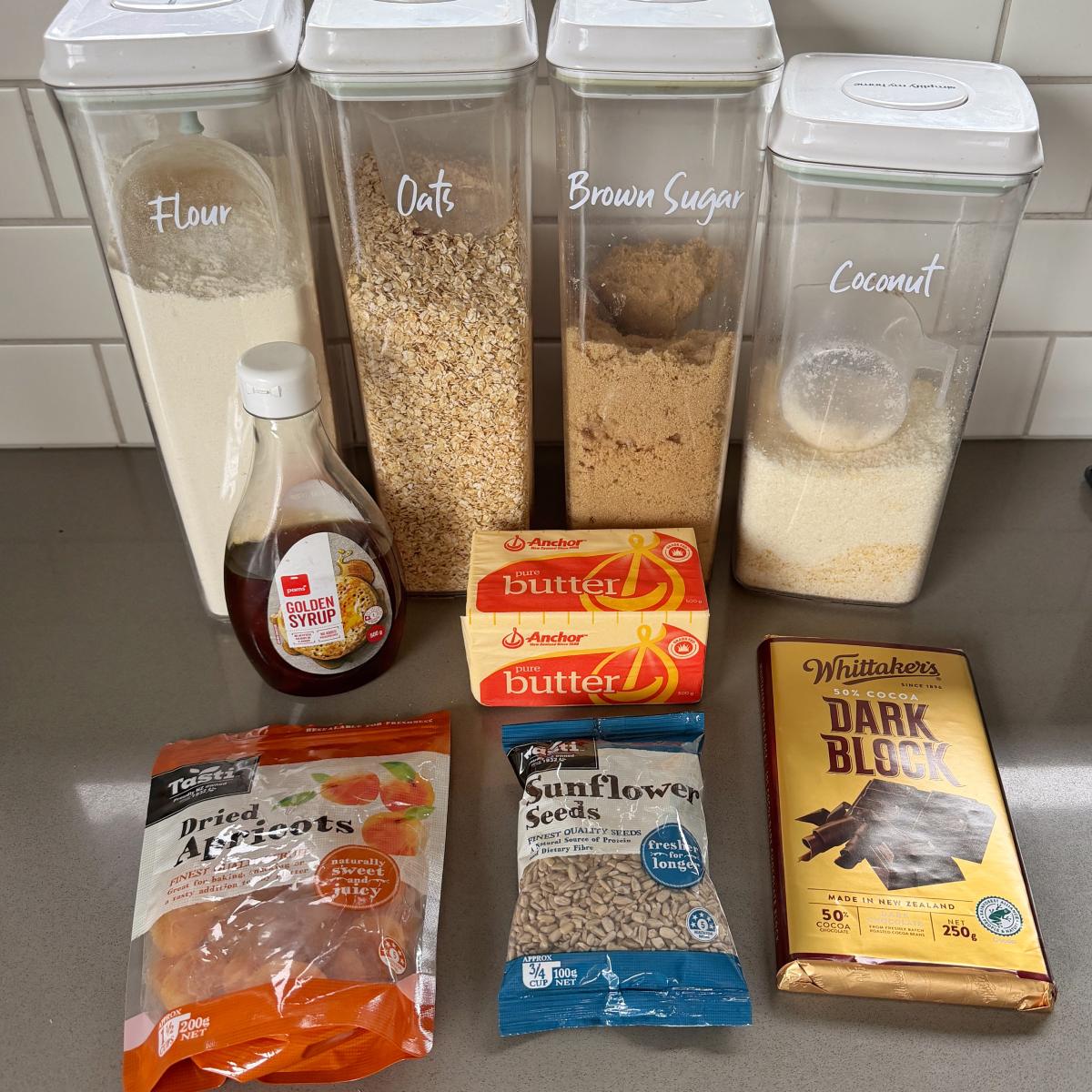 Apricot Bumper Bars Baking ingredients on a counter: containers of flour, oats, brown sugar, coconut; syrup, butter, dried apricots, sunflower seeds, and a block of dark chocolate.