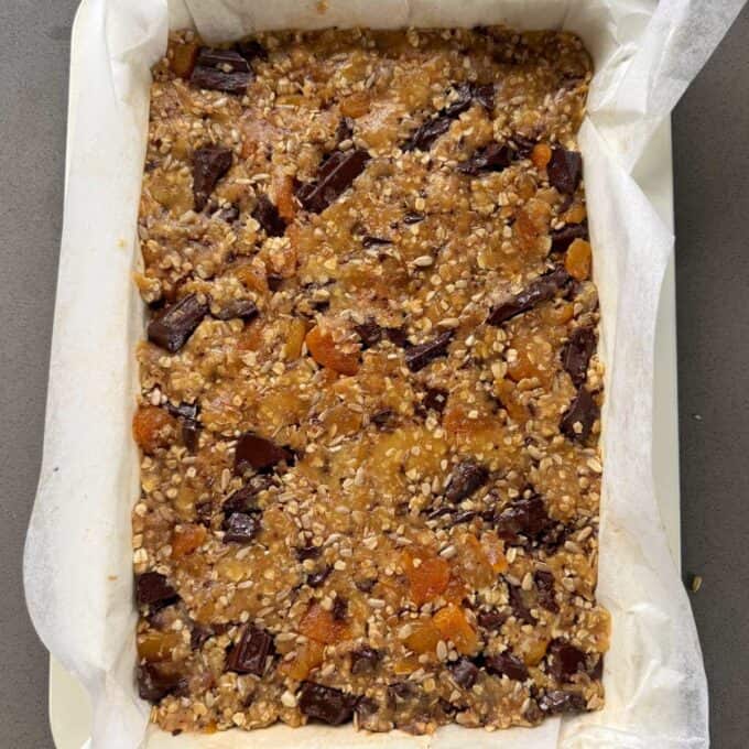 Apricot Bumper Bars A rectangular baking dish lined with parchment paper contains an unbaked mixture of oats, dried fruit, and chocolate chunks.