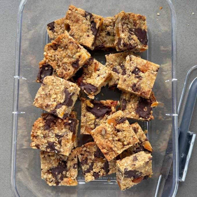 Apricot Bumper Bars A clear container holds several square oat and chocolate chip cookie bars; a serving utensil is placed to the right inside the container.