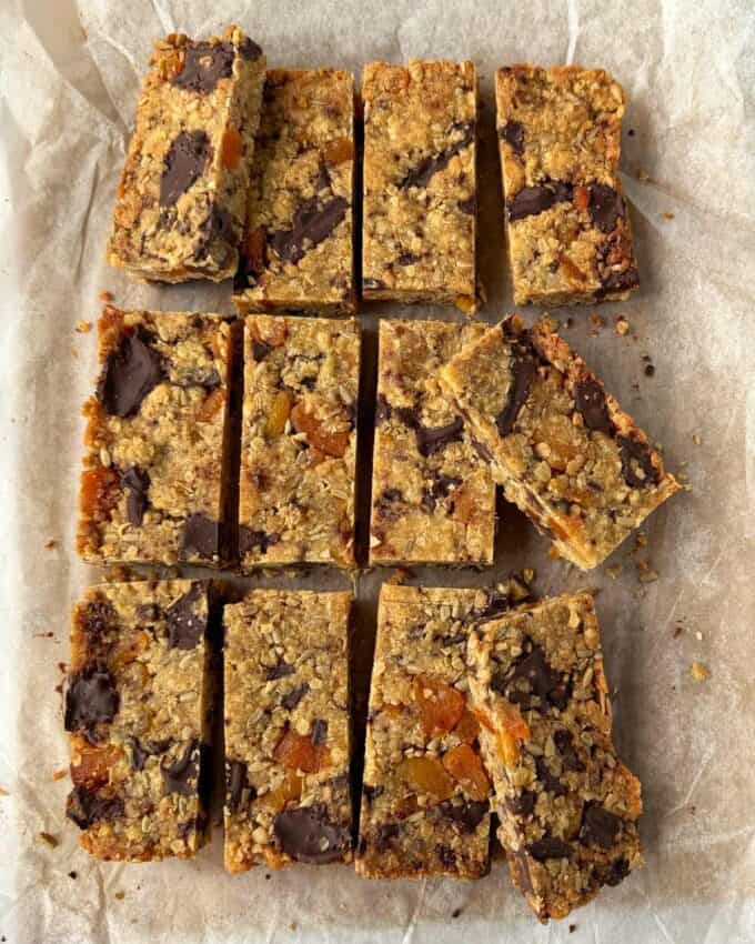 Apricot Bumper Bars Thirteen rectangular oat bars with chocolate chunks and fruit pieces are arranged on parchment paper, some slightly separated or overlapping.