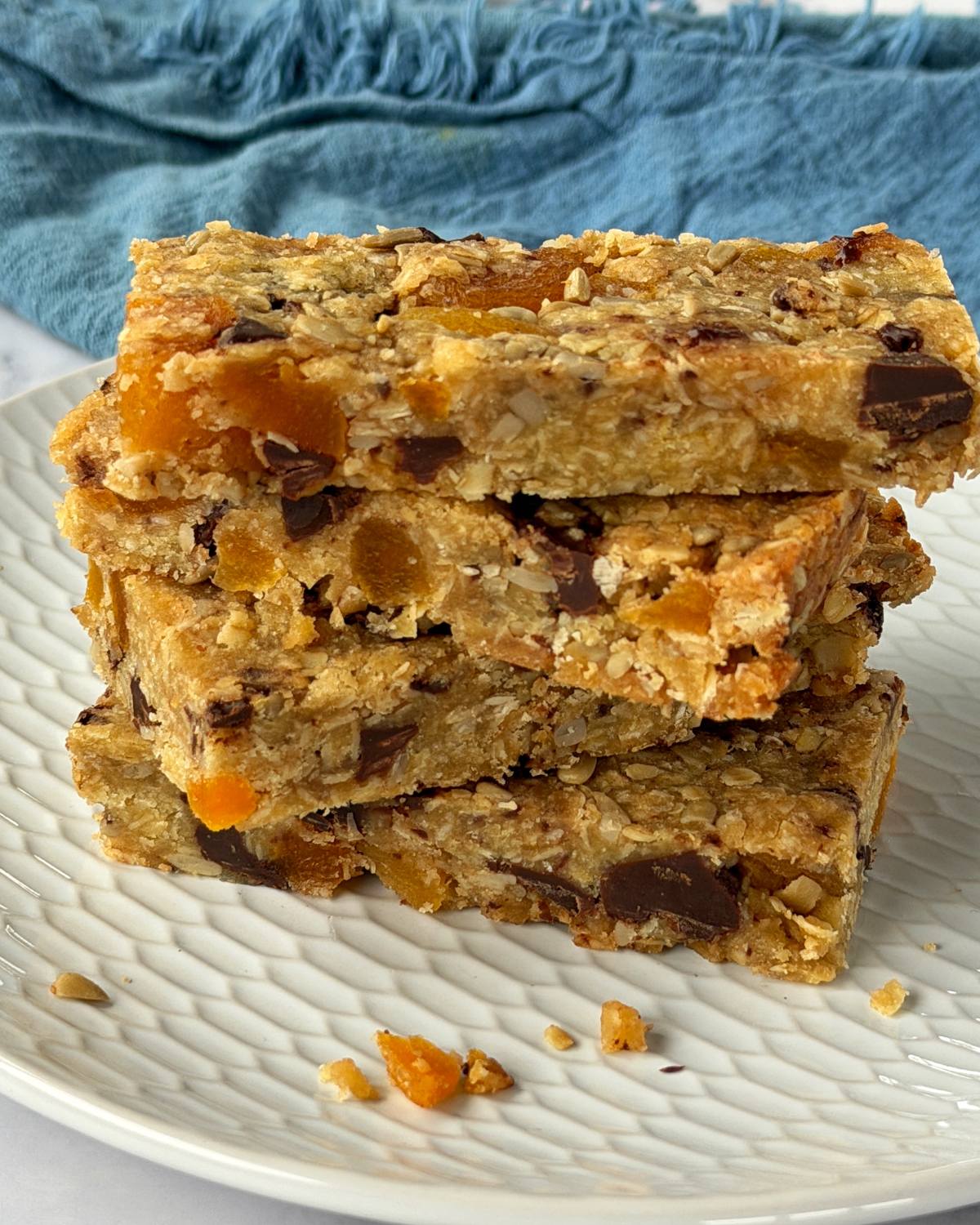 Apricot Bumper Bars A stack of four oat and fruit granola bars with visible chocolate chunks, placed on a white plate with a blue cloth in the background.