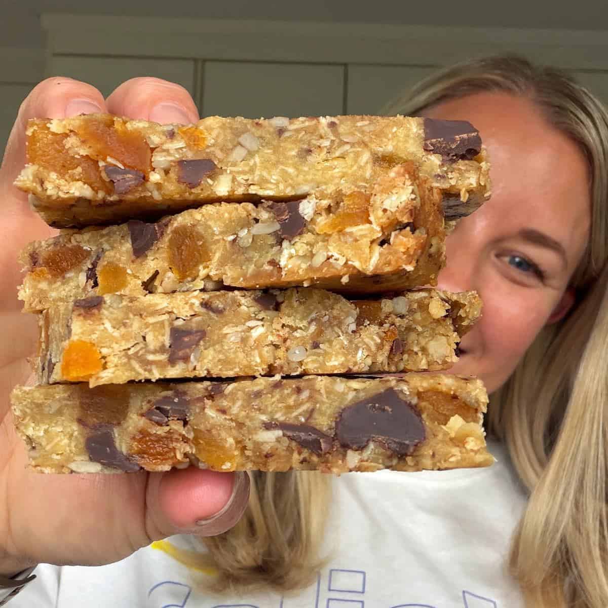 Apricot Bumper Bars A person holds up a stack of four homemade granola bars with visible chocolate chunks and dried fruit pieces, in a kitchen setting.