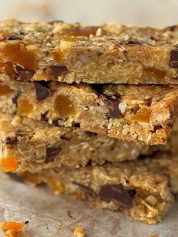 A stack of four homemade granola bars with visible chunks of dried fruit and chocolate on a parchment-lined surface.