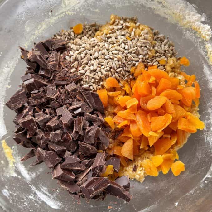 Apricot Bumper Bars Chopped dark chocolate, dried apricots, and sunflower seeds in a glass mixing bowl with oat mixture.