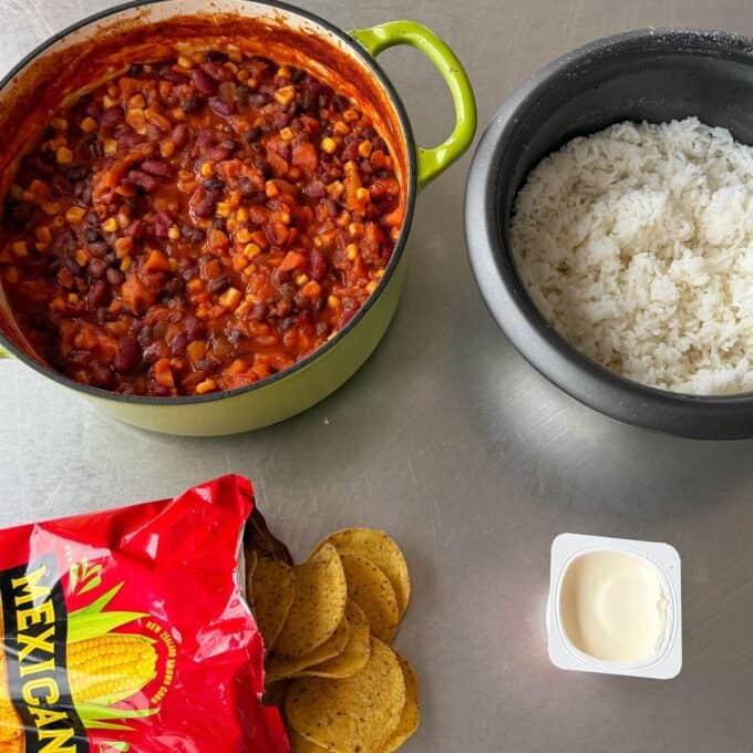 Baked Vegetarian Chilli A pot of bean chili, a rice cooker with white rice, a bag of tortilla chips, some chips on the table, and a container of sour cream are arranged on a metal surface.