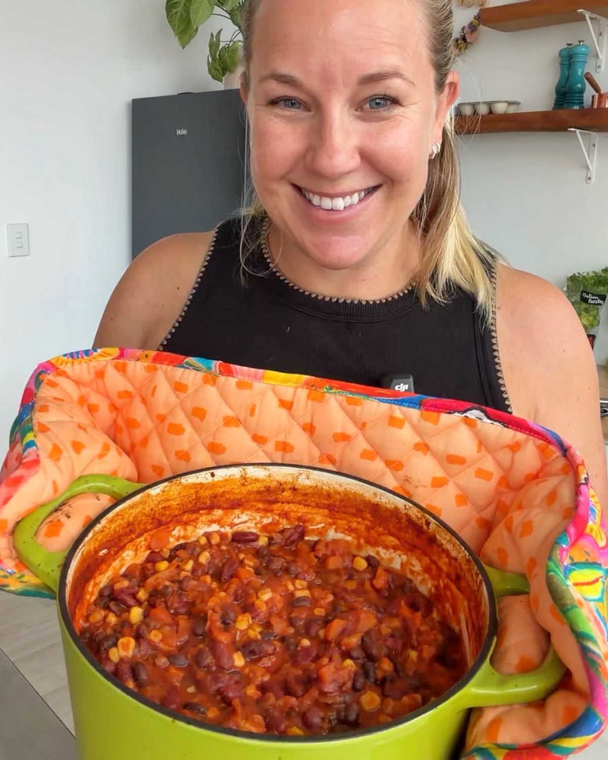 Baked Vegetarian Chilli A woman holds a green pot filled with chili containing beans, corn, and tomato sauce, smiling at the camera while wearing an oven mitt.