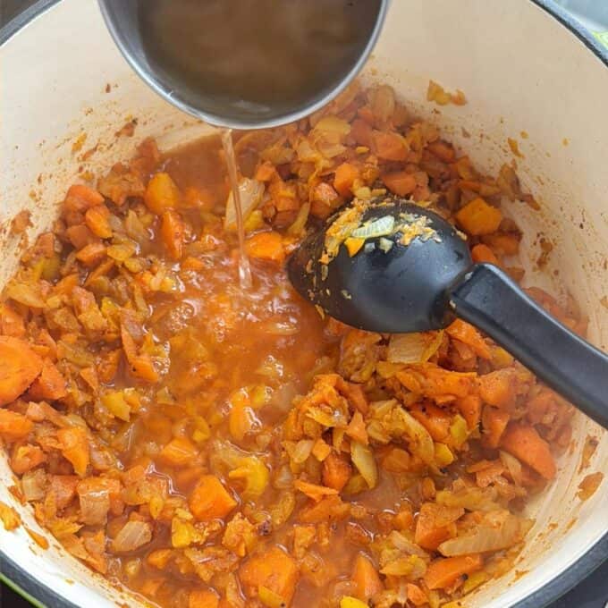 Baked Vegetarian Chilli A pot of sautéed chopped onions and carrots being deglazed with liquid poured from a small metal container, with a black cooking spatula in the mixture.