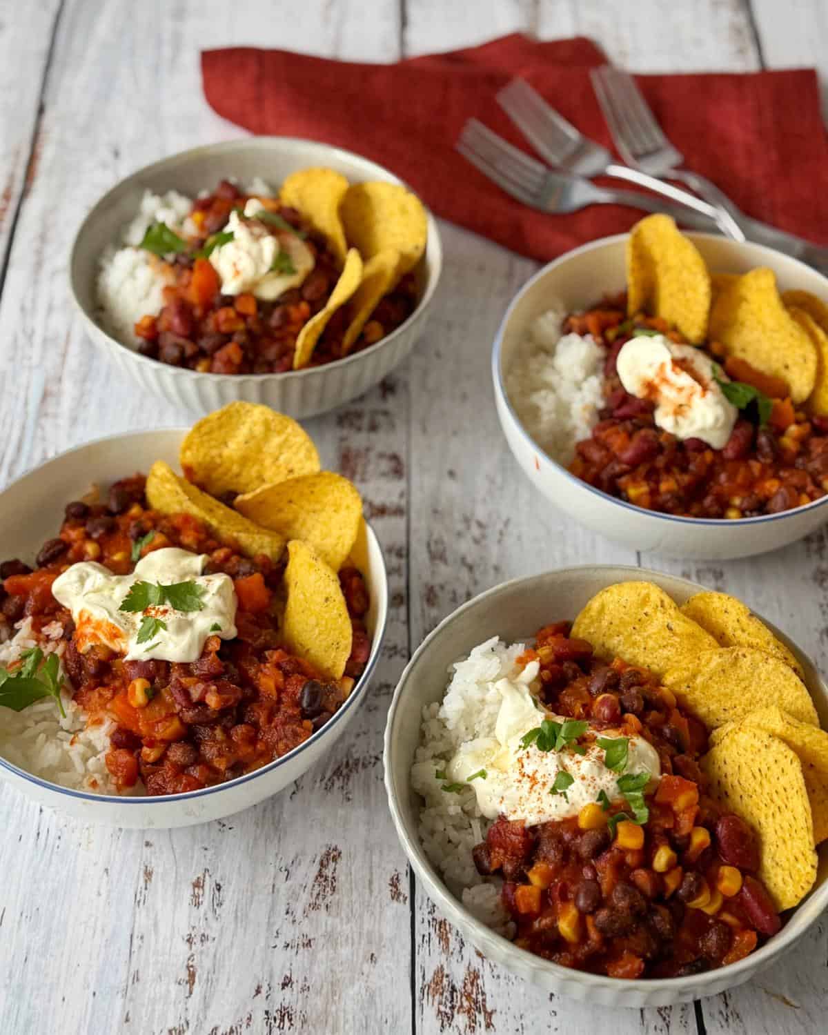 Baked Vegetarian Chilli Four bowls of chili with rice, corn chips, and sour cream are arranged on a white wooden table with a red cloth and three forks in the background.