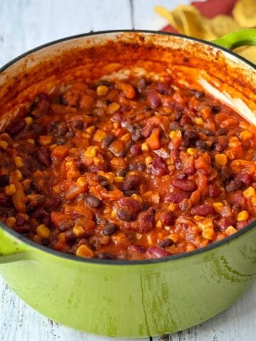 A green pot filled with vegetarian chili containing beans, corn, and tomatoes on a white wooden surface.
