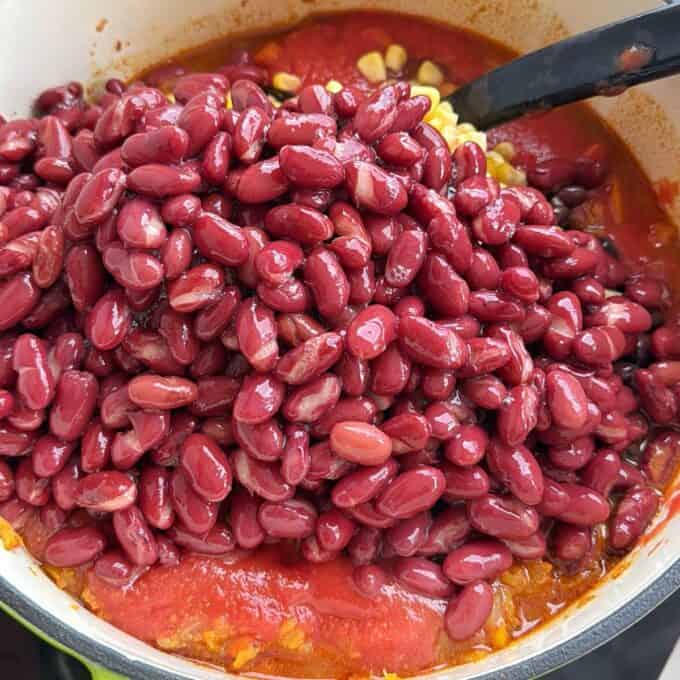 Baked Vegetarian Chilli A close-up of red kidney beans on top of tomato sauce and corn in a pot, with a black spoon partially visible.