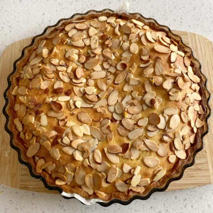 Bakewell Tart A round baked tart topped with sliced almonds sits on a wooden cutting board atop a light-colored countertop.