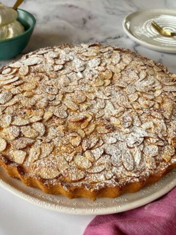 A round almond tart topped with sliced almonds and powdered sugar sits on a white plate, with a bowl of whipped cream and a dessert plate in the background.