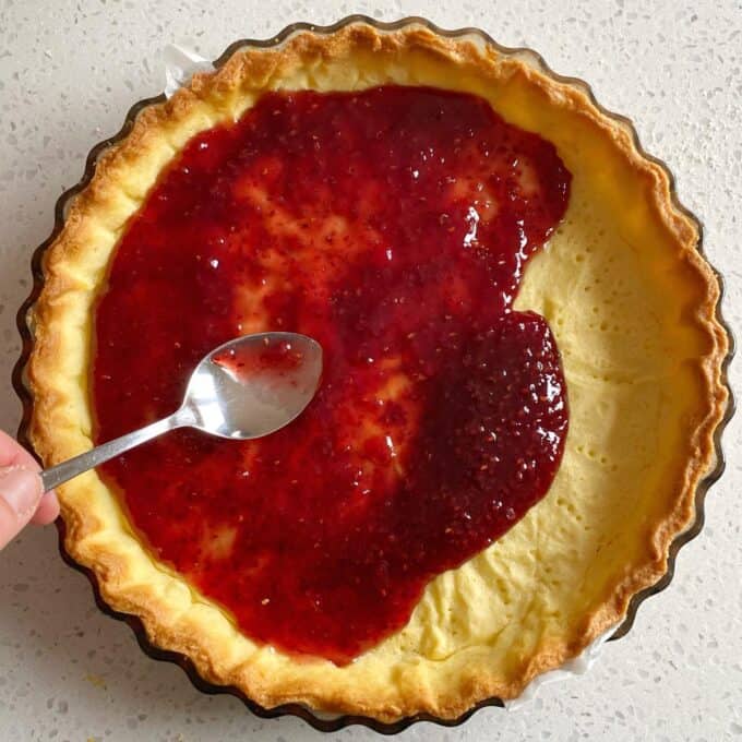 Bakewell Tart A hand spreads red raspberry jam onto a partially filled pastry tart crust using a spoon.