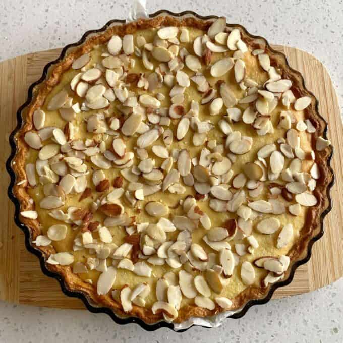 Bakewell Tart A baked tart topped with sliced almonds sits on a round wooden cutting board, placed on a speckled countertop.