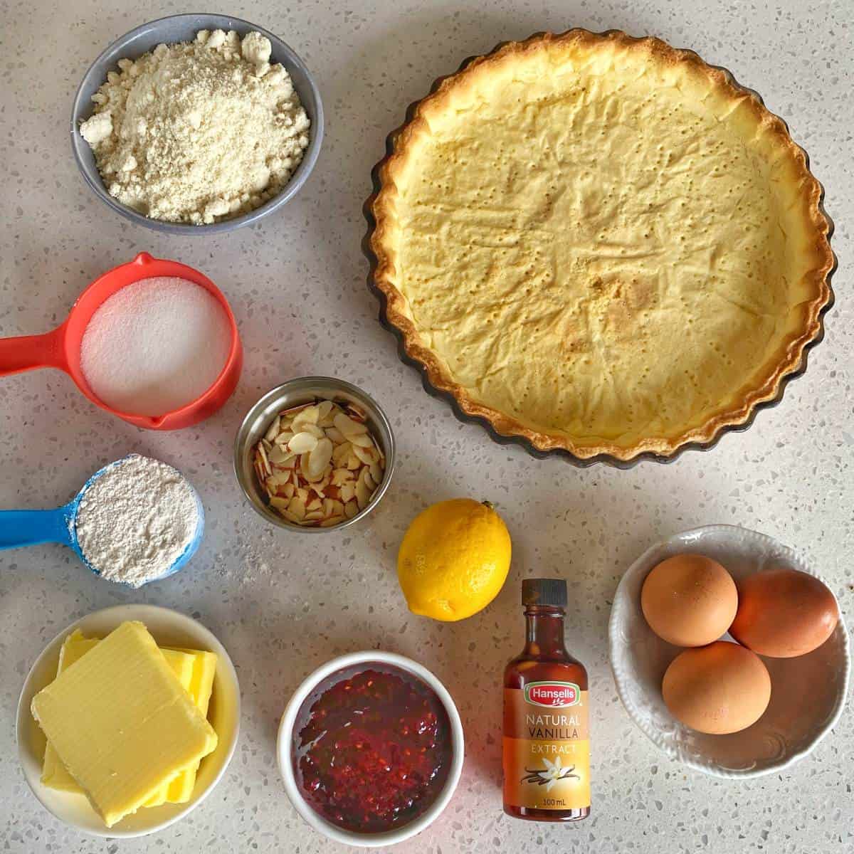 Bakewell Tart Baked tart crust in a pan surrounded by ingredients: flour, sugar, sliced almonds, lemon, vanilla extract, eggs, butter, and a bowl of red jam on a countertop.