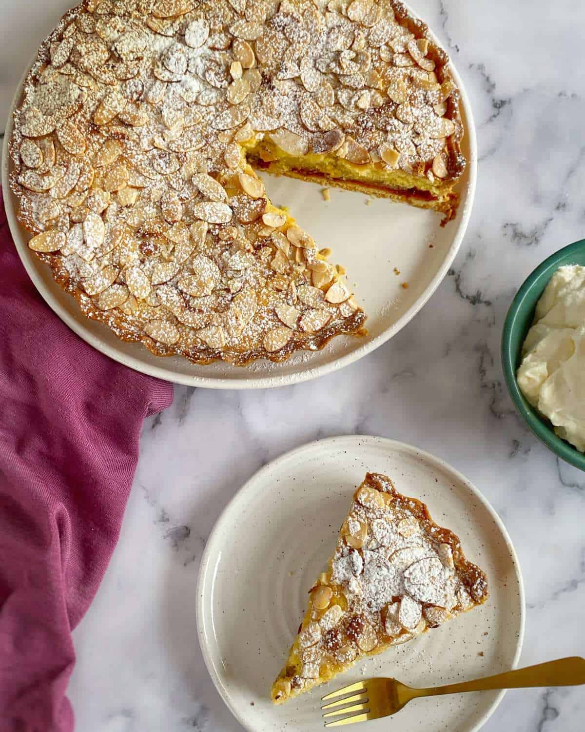 Bakewell Tart A round almond cake with a slice missing, dusted with powdered sugar. The slice is served on a plate with a fork. A bowl of whipped cream and a pink cloth are nearby.