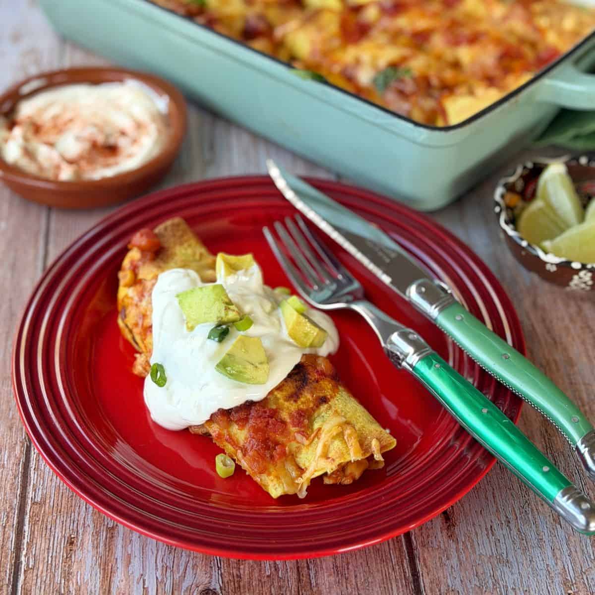 Cheesy Chicken Enchiladas A plate with two enchiladas topped with sour cream and avocado, with a fork and knife on the side. In the background are a dish of sour cream, lime wedges, and a baking dish of enchiladas.