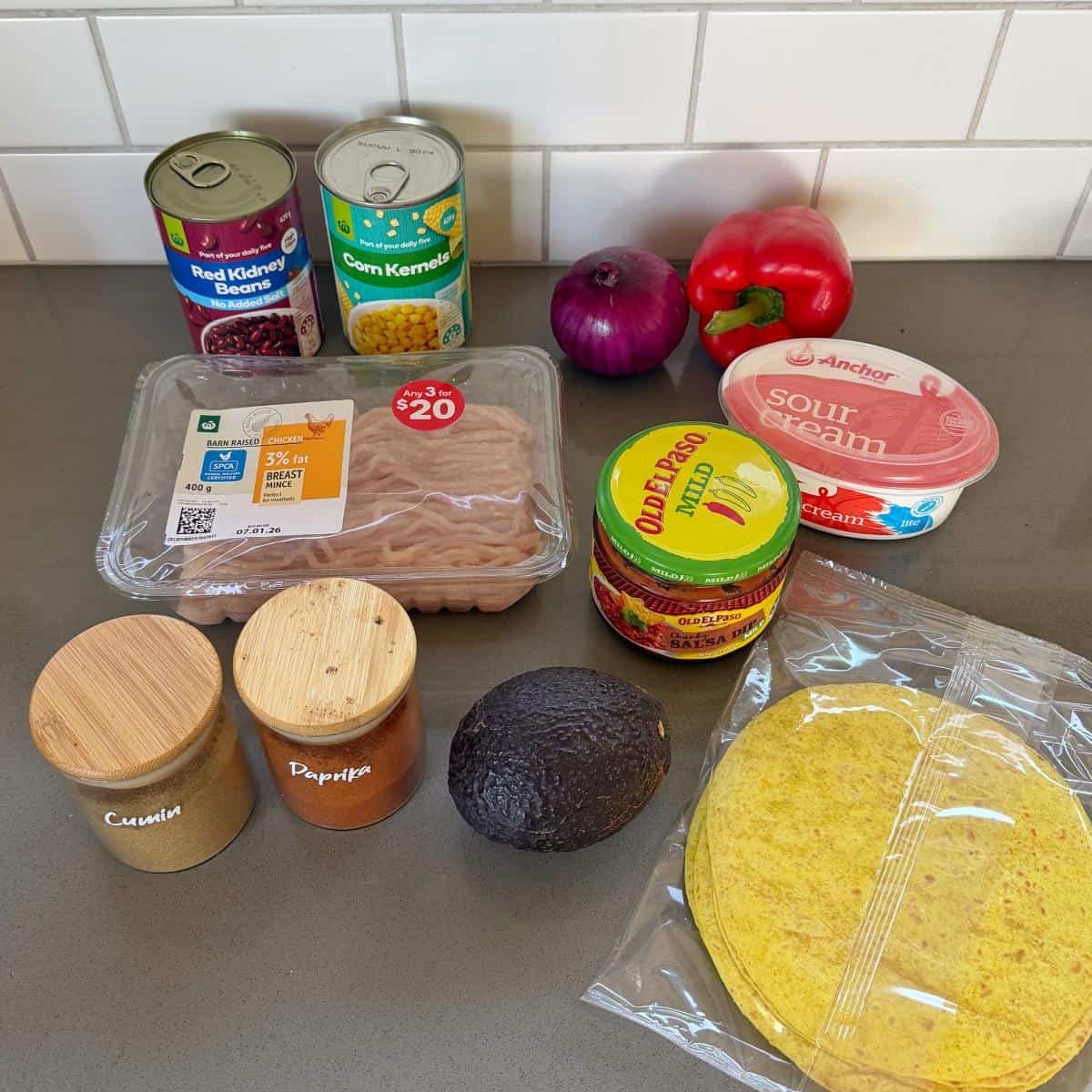 Cheesy Chicken Enchiladas A selection of taco ingredients on a countertop, including ground chicken, canned beans, corn, red onion, red pepper, sour cream, taco shells, avocado, taco sauce, cumin, and paprika.