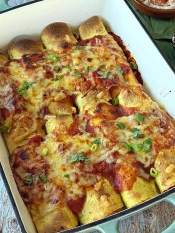A baking dish filled with baked enchiladas topped with melted cheese, red sauce, and chopped green onions sits on a wooden table.