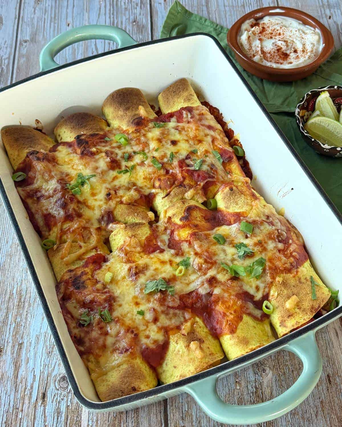 Cheesy Chicken Enchiladas A baking dish filled with cheesy enchiladas topped with sauce, garnished with herbs, next to bowls of sour cream and lime wedges.