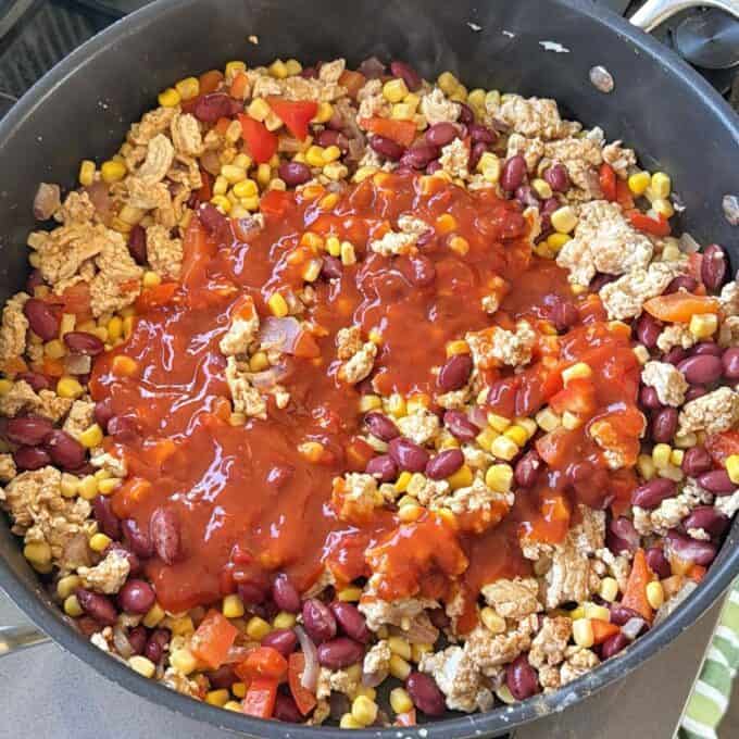 Cheesy Chicken Enchiladas A large pan filled with cooked ground meat, kidney beans, corn, chopped red bell peppers, and topped with tomato sauce.