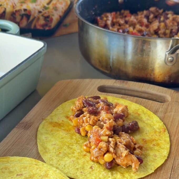 Cheesy Chicken Enchiladas A yellow tortilla on a wooden board topped with a mixture of beans, corn, and meat, with a pot of the same filling and a tray of rolled tortillas in the background.