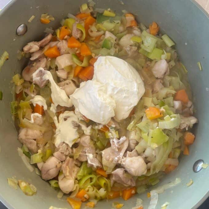 Chicken and Leek Pie Chopped chicken, carrots, and leeks are being cooked in a pot with a large dollop of cream or mayonnaise added on top.