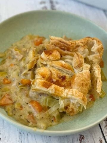 A serving of creamy vegetable pot pie with a flaky pastry crust in a light green bowl on a white wooden surface.