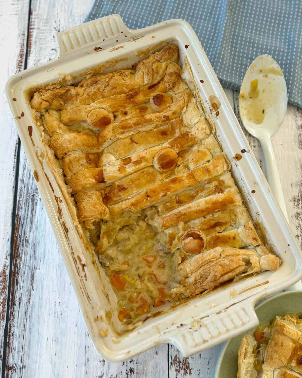 Chicken and Leek Pie Rectangular baking dish with a partially eaten puff pastry-topped casserole, showing filling with vegetables; a serving spoon with residue is on the side.