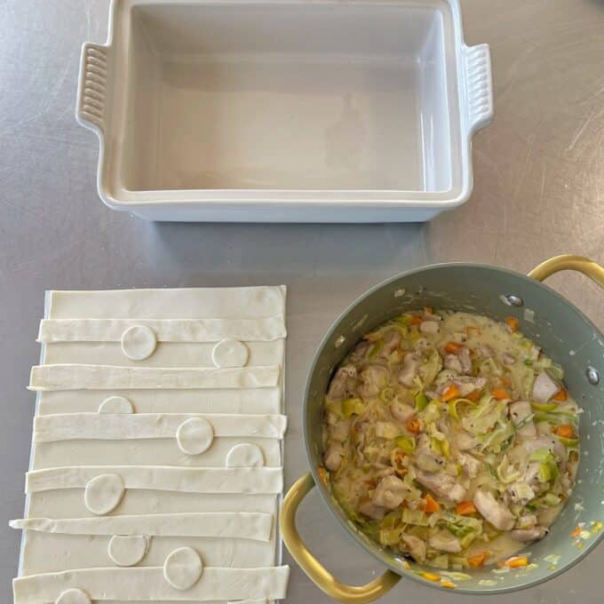 Chicken and Leek Pie A baking dish, strips of pastry dough, and a pot filled with a creamy chicken and vegetable mixture are arranged on a countertop.