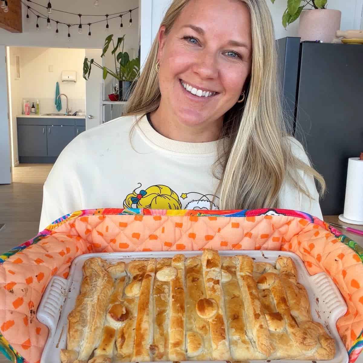 Chicken and Leek Pie A woman smiles while holding a rectangular baked pastry in a ceramic dish lined with an orange-patterned cloth.
